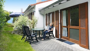 Une terrasse avec une table et des chaises, une personne assise, et une vue sur un bâtiment doté de grandes fenêtres et de volets.