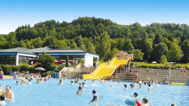 Une piscine publique avec un toboggan jaune et une colline boisée en arrière-plan.