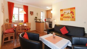 A cozy living room with a black sofa, red cushions, a dining area with wooden chairs, a small kitchen, and a dining table with a checkered tablecloth. - Ferienwohnung in Bodenmais am Grossen Arber (Bodenmais)