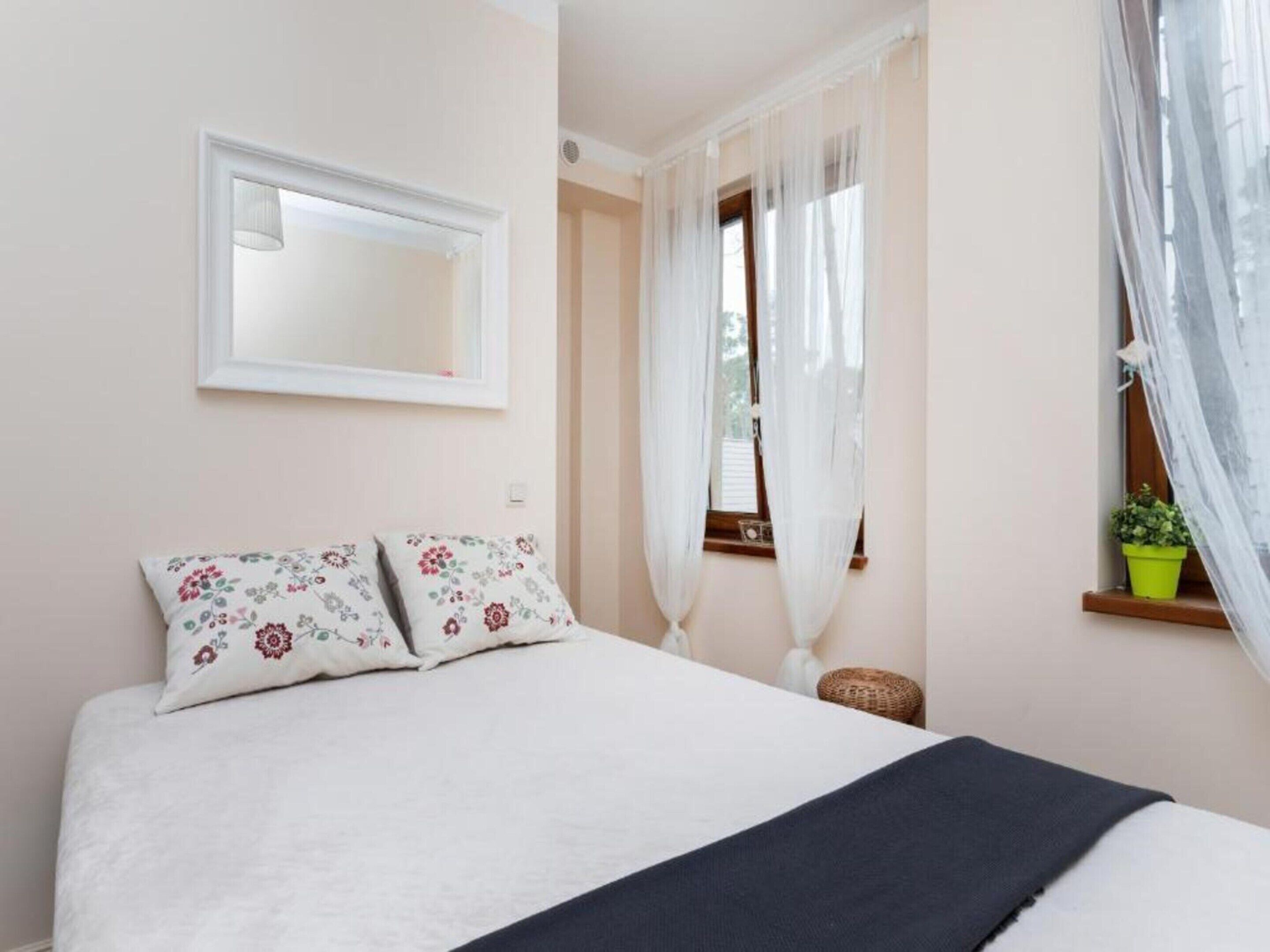 Une chambre à coucher avec un lit, une fenêtre ornée de rideaux légers, une petite plante en pot et un miroir accroché au mur.