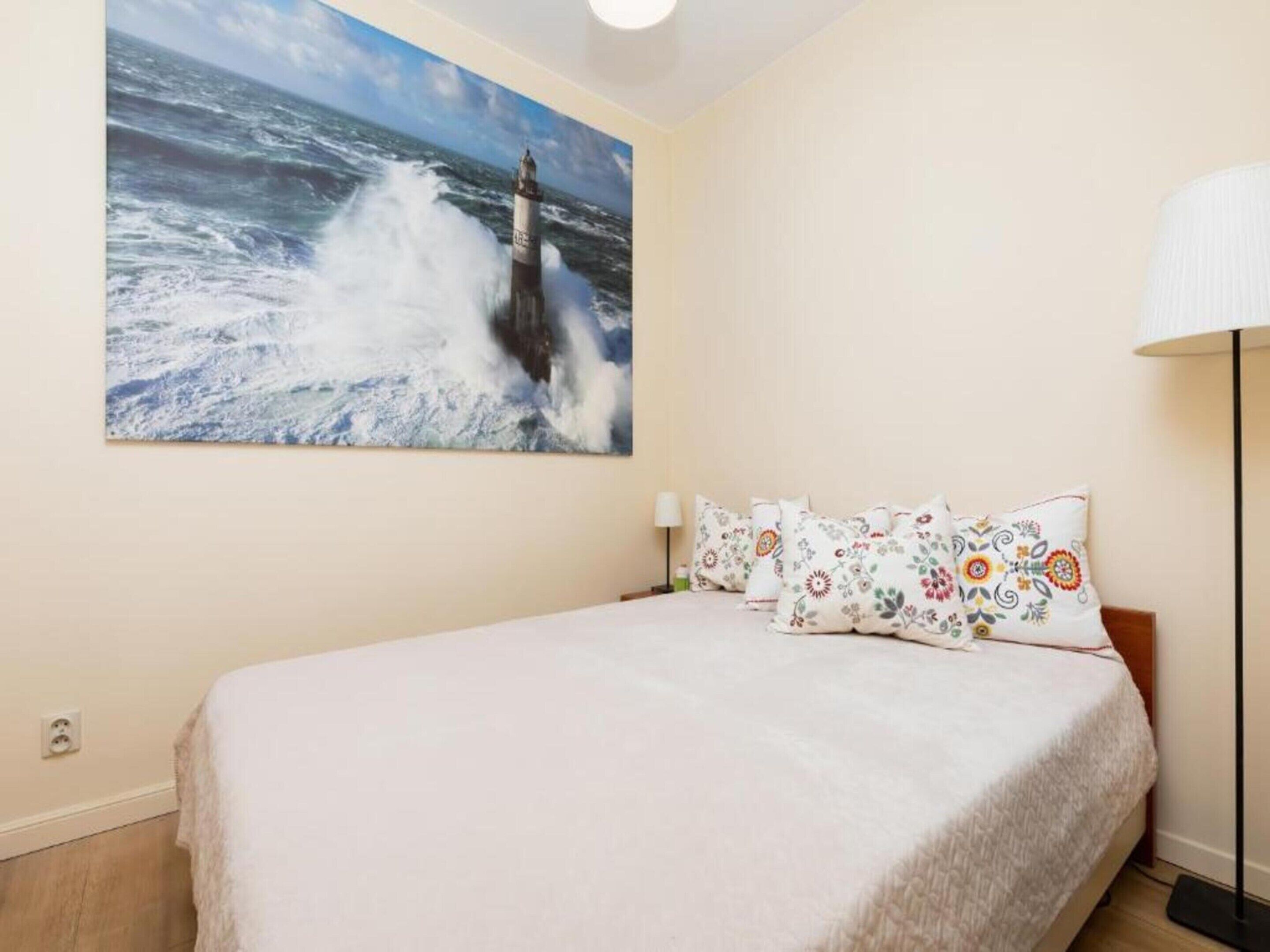 Une chambre à coucher avec un grand tableau représentant un phare au mur, un lit recouvert d’une courtepointe blanche et des oreillers à motifs floraux.