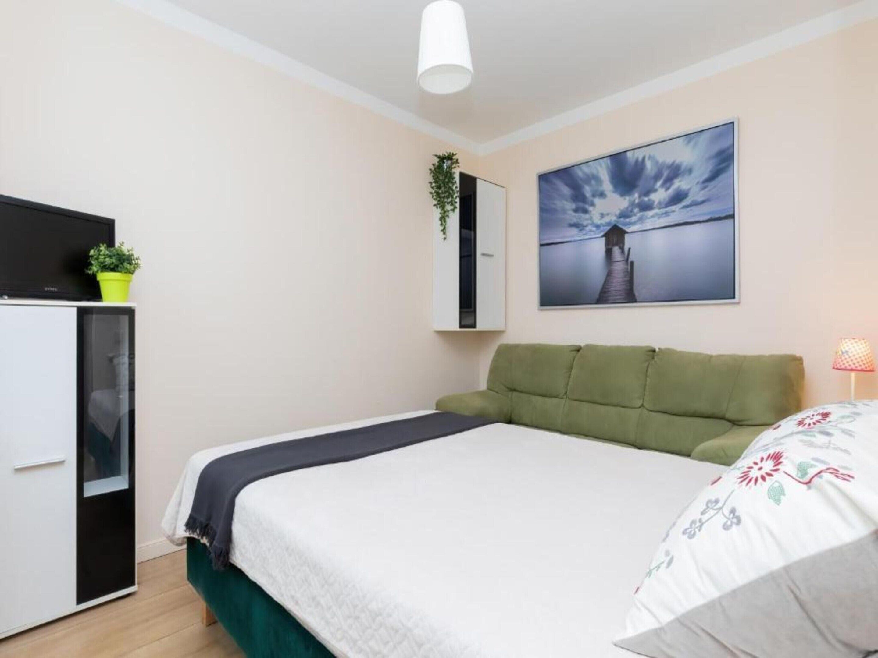 A bedroom with a bed, a desk, a chair, a potted plant, and a painting on the wall.