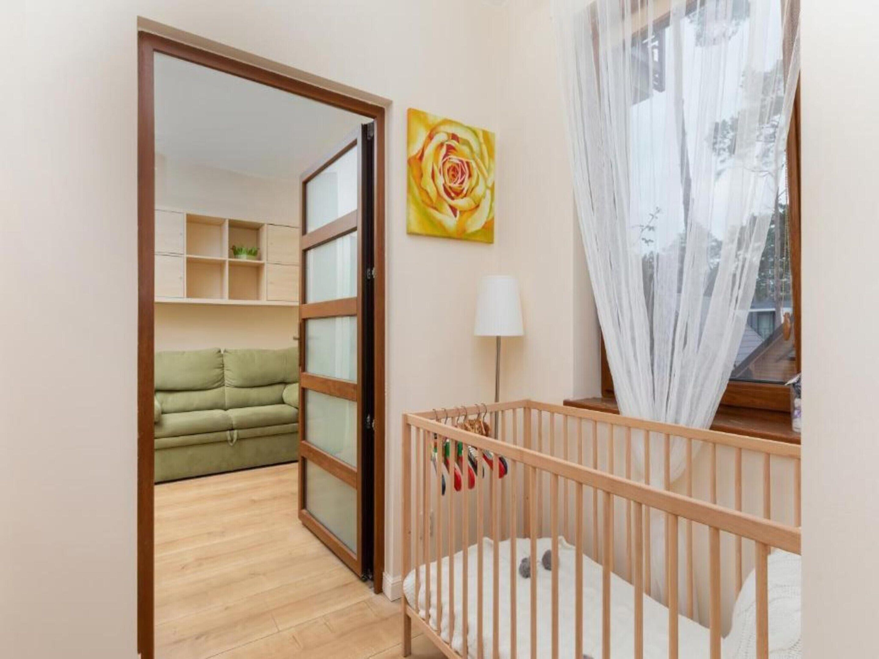 Ein Zimmer mit einem hölzernen Wiege, einem grünen Sofa, einem Wandbild und einer Glasschiebetür, die zu einem weiteren Raum mit einem weißen Sofa und Regalen führt.