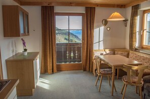 A room with a wooden desk, a dining table with chairs, a bench, a window with curtains, and a view of a mountainous landscape.