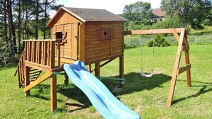 A wooden playhouse with a slide and swing set in a grassy yard.