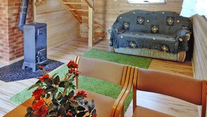 A cozy cabin interior with a wood-burning stove, a sofa, a dining table with a vase of flowers, and a staircase.