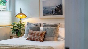 Une chambre à coucher avec un lit, un tableau encadré représentant un paysage côtier, une lampe et une plante en pot.