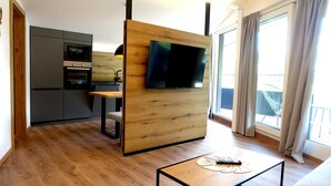 A modern living room with a wooden partition, a flat-screen TV, a small table with a remote control, and a kitchen in the background.