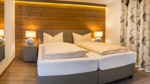 Un lit double avec une tête de lit grise et du linge de lit blanc, deux tables de chevet avec des lampes et un panneau mural en bois.