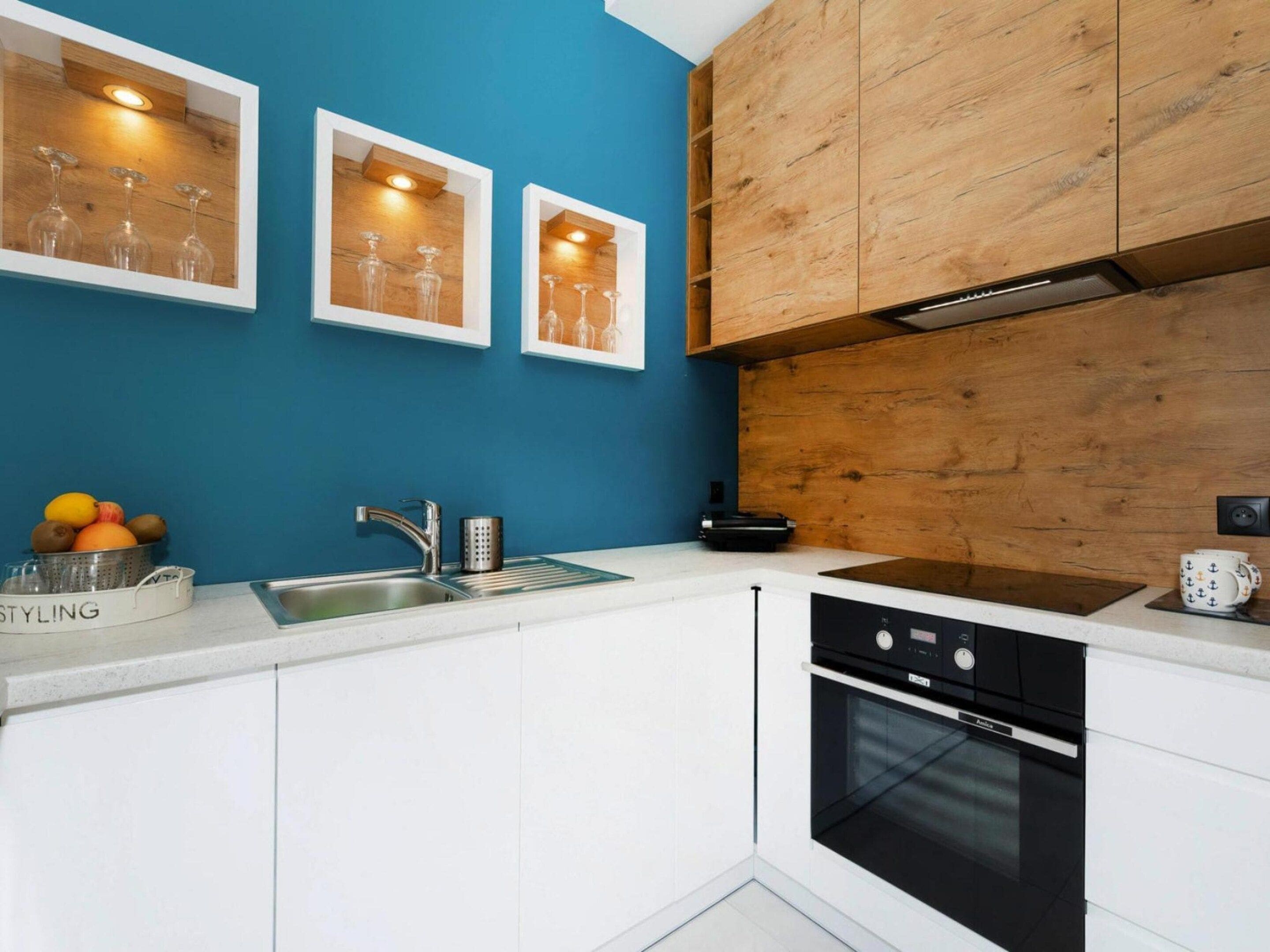 A kitchen with white cabinets, a blue wall, wooden upper cabinets, a sink, a stove, and framed pictures on the wall.