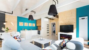 A modern living room with white sofas, a coffee table, and a fireplace. There is a kitchen area with wooden cabinets and a TV mounted on the wall.