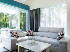 A modern living room with a grey sofa, colorful cushions, a white coffee table, and large windows with curtains. - Holiday Home in Ustronie Morskie Near Beach (Ustronie Morskie)