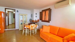 Una sala de estar con un sofá naranja, una mesa de comedor con sillas y una cocina visible a través de un umbral abierto.