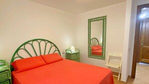 Un dormitorio con estructura de cama metálica verde, colcha roja, mesita de noche verde con lámpara, cómoda verde, una silla blanca y un espejo en la pared.
