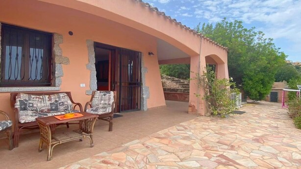 Un edificio con un patio de baldosas de piedra, una zona de estar cubierta con dos sillas y una mesa, y un ventanal con cortinas.