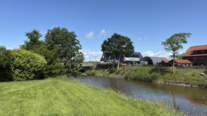 A riverside view with a grassy bank, a bridge, and buildings in the background. - Ferienwohnung, Fedderwardersiel (Butjadingen)