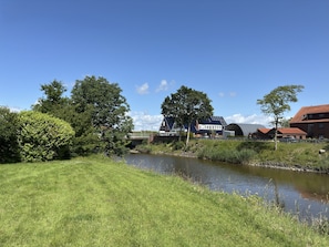 A riverside view with a grassy bank, a bridge, and buildings in the background. - Ferienwohnung, Fedderwardersiel (Butjadingen)