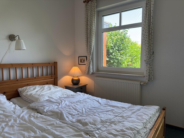 A bedroom with a wooden bed, a bedside lamp, a window with curtains, and a radiator. - Ferienwohnung, Fedderwardersiel (Butjadingen)