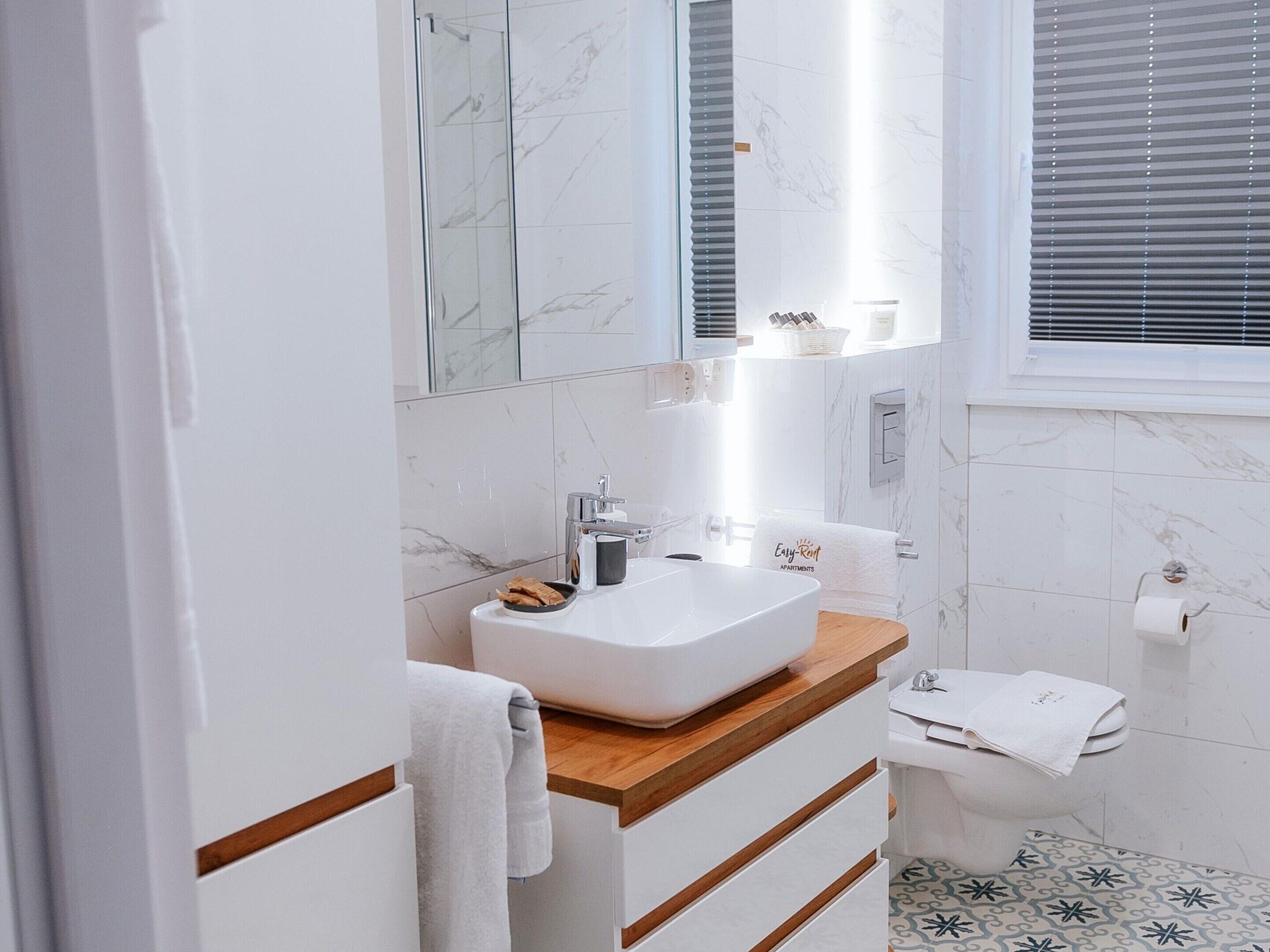 Een moderne badkamer met een witte wastafel, een houten opbergmeubel en een betegelde muur van marmer.