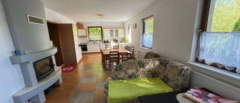 Ein Wohnzimmer mit einer Couch, einem Tisch und einem Kamin. Im Hintergrund ist eine Küche zu sehen.