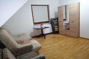 Una stanza piccola con pavimento in legno, un divano, un termosifone, una finestra con persiana decorata e uno specchio a parete.