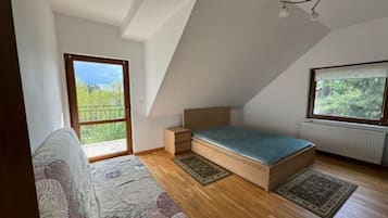 A small, loft-style bedroom with a sloped ceiling, a single bed, a nightstand, a window with a view of greenery, and a door leading to an outdoor balcony.