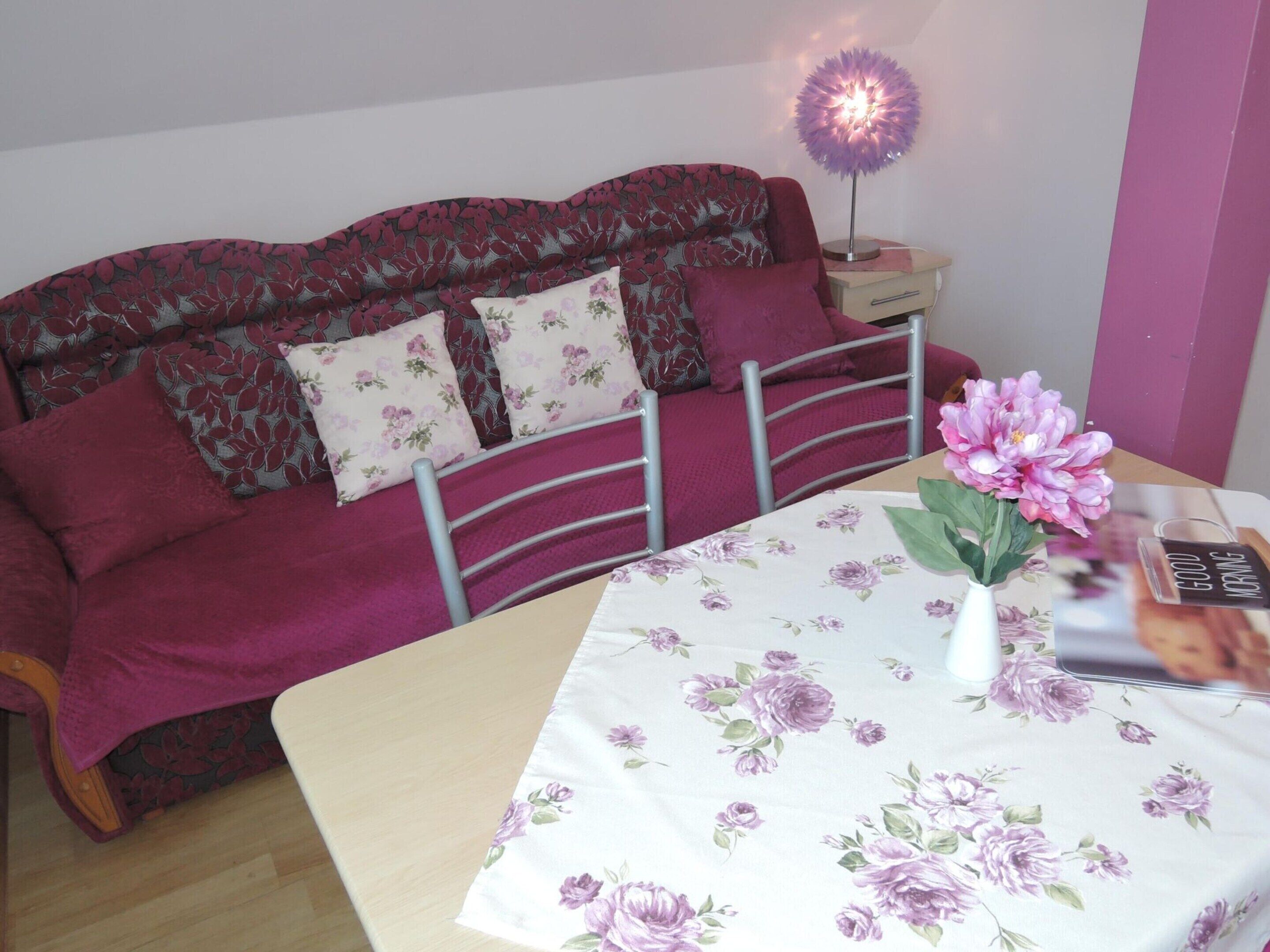 A cozy bedroom with a bed, a bedside table with a lamp, a floral tablecloth, and a vase with flowers.