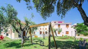Um parquinho com balanços e escorrega, cercado por grama e árvores, com edifícios ao fundo.