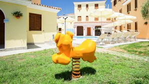 Um hidrante amarelo numa área gramada, com uma piscina e guarda-sóis ao fundo.