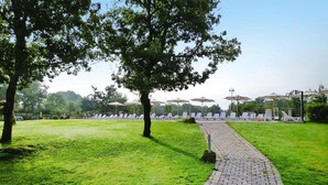 Un chemin pavé mène à un espace aménagé avec des sièges et des parasols, entouré de pelouse et d’arbres.
