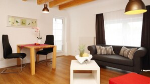 A modern living room with a wooden floor, a black sofa with striped cushions, a white coffee table, and a red ottoman. There is a dining table with chairs and a vase of flowers. - Apartment in Eifel With Terrace and Nature (Lissendorf)