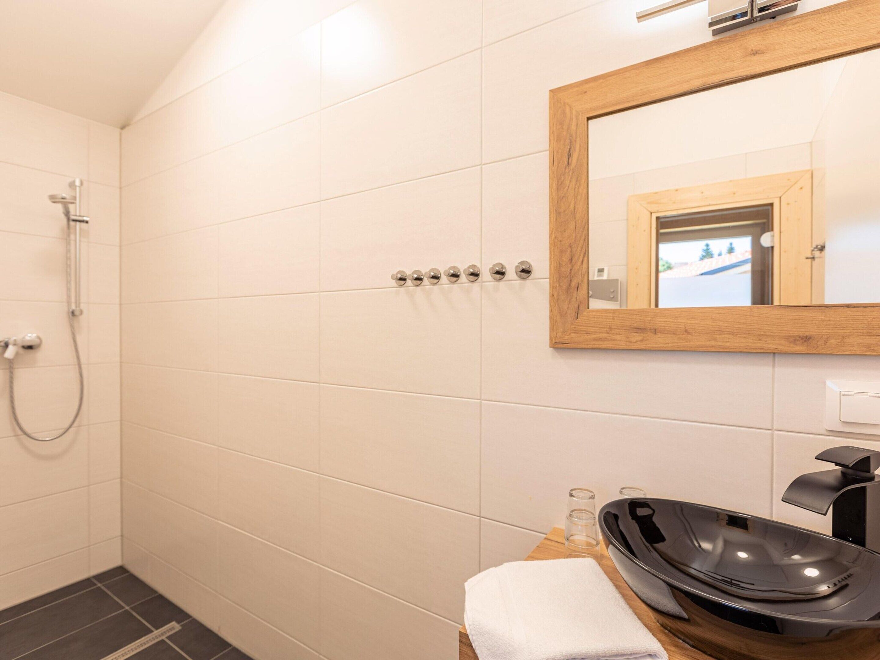 Un bagno con piastrelle bianche, uno specchio con cornice in legno, un lavandino nero e un soffione a mano.