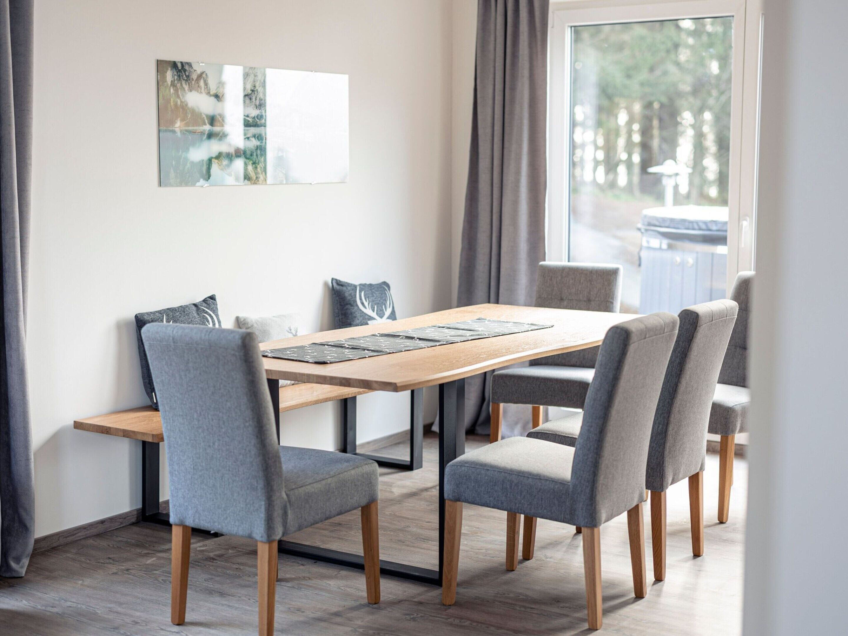 Un espace repas moderne avec une table en bois, des chaises à revêtement gris et une grande fenêtre avec des rideaux.
