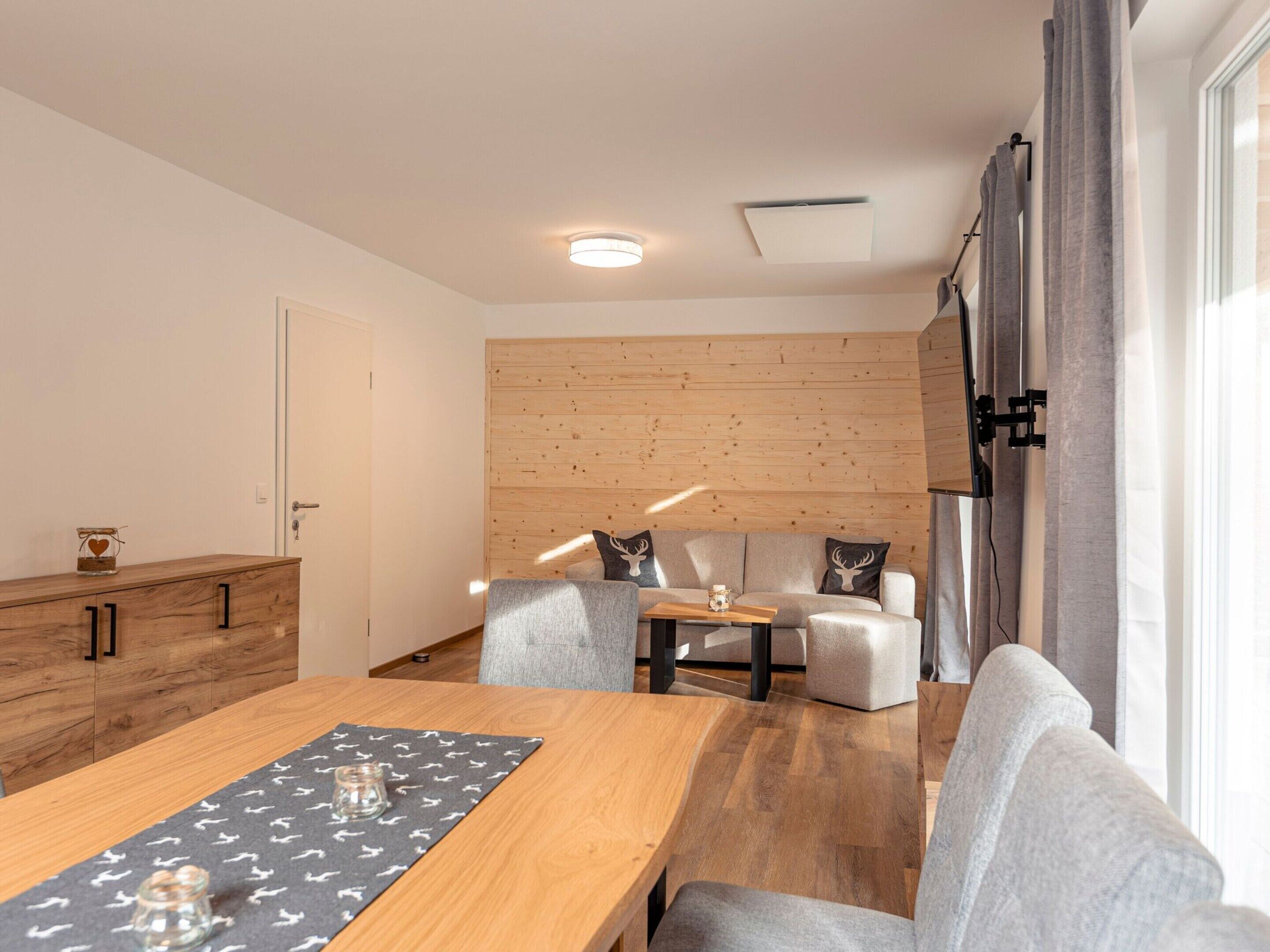 A modern living room with a wooden dining table, grey sofas, and a wooden accent wall.