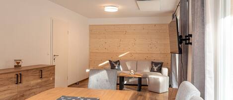 A modern living room with a wooden dining table, grey sofas, and a wooden accent wall.