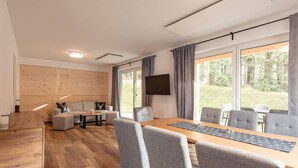 A modern living room with a wooden floor, grey sofas, a wooden coffee table, and a large window with curtains. - Chalet Arber in St.englmar With its own Hottube (Sankt Englmar)