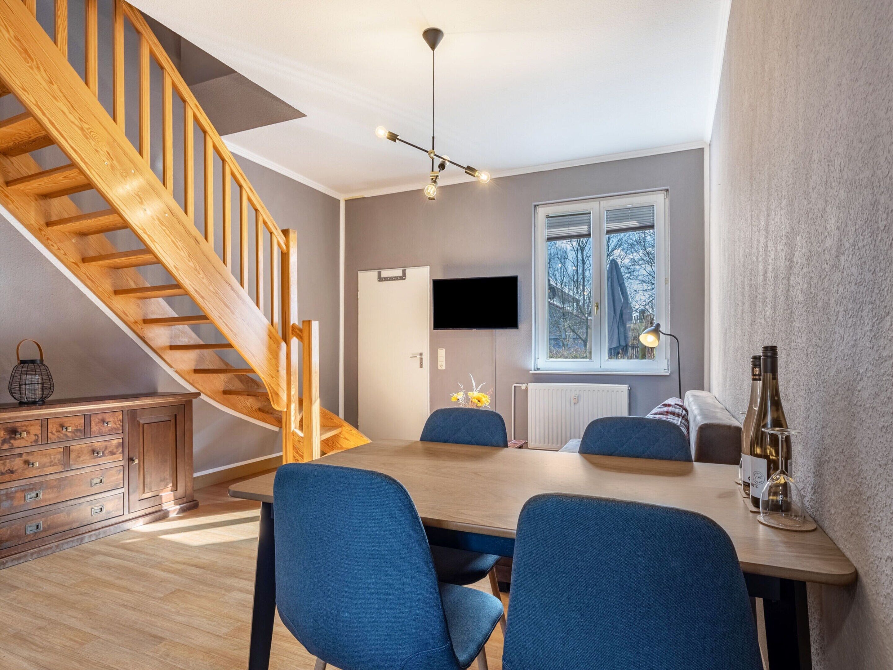 A modern living room with a wooden staircase, a dining table with chairs, a TV mounted on the wall, and a window with a view of trees.