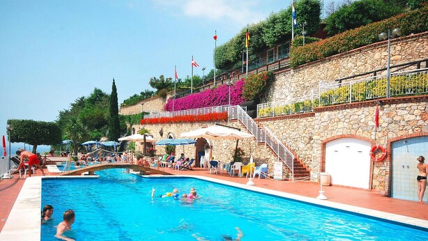 A swimming pool with people in the water, surrounded by a stone wall, and a staircase leading to a balcony with plants and flags. - Holiday Flat in Pretty Residence With Communal Pool in Pietra Ligure (Pietra Ligure)