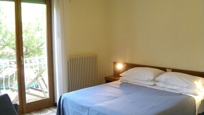 Une chambre à coucher comprenant un lit, une table de chevet, une fenêtre donnant sur une terrasse extérieure et un radiateur.