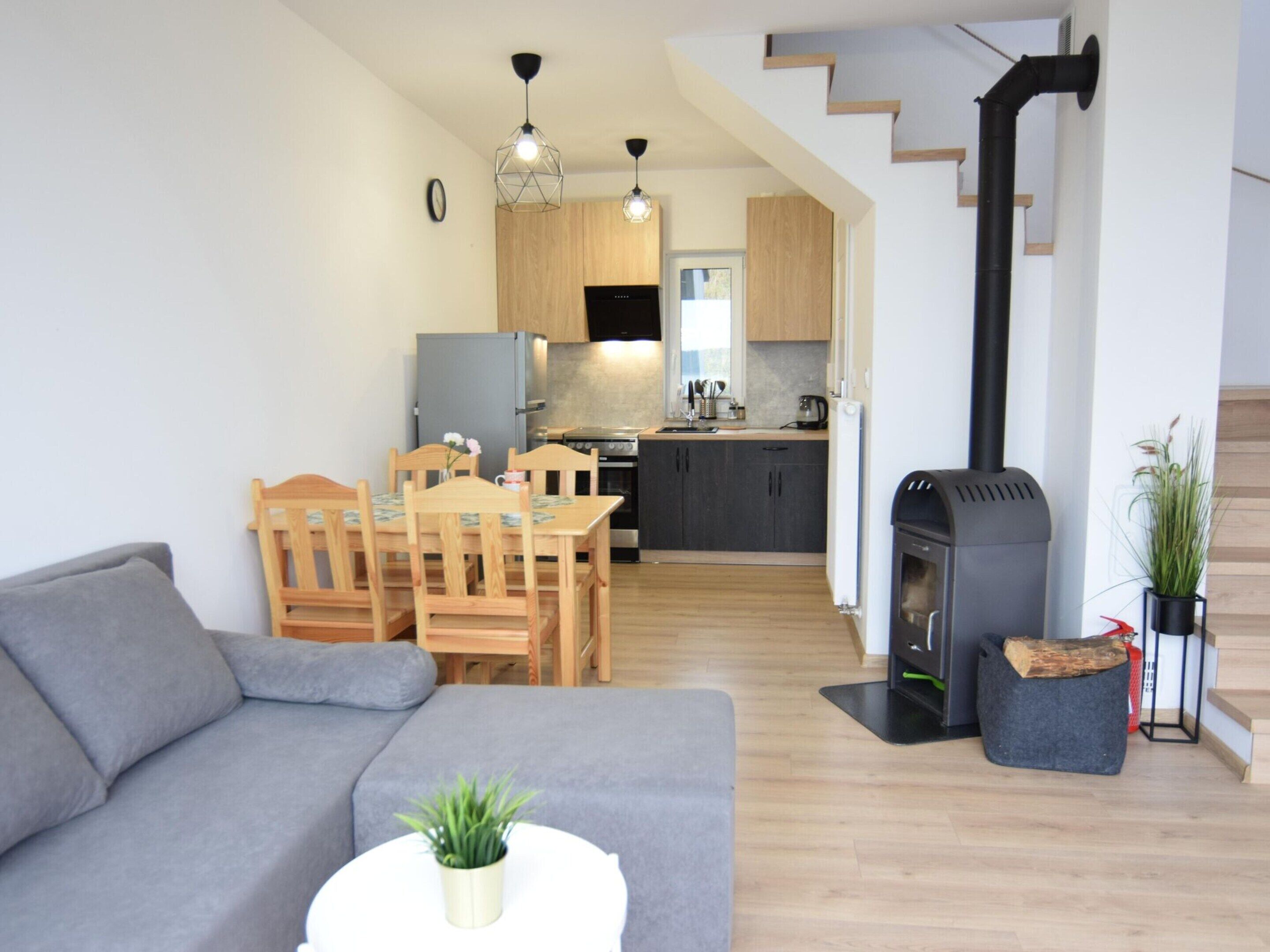 A modern living room with a grey sofa, wooden dining table, and chairs. A kitchen with wooden cabinets and a black stove is visible in the background.