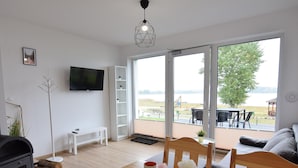 Ein modernes Wohnzimmer mit einem Holzesstisch, einer Couch und einem an der Wand befestigten Fernseher. Ein großes Fenster bietet einen Blick ins Freie.