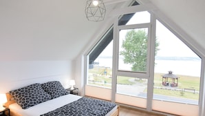 Ein Schlafzimmer mit einem Bett, einem Nachttisch, einer Lampe und Blick auf einen Park durch große Fenster.