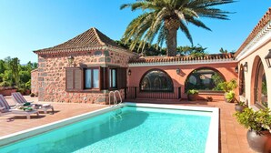 Une piscine avec des chaises longues, un bâtiment en pierre et un palmier.
