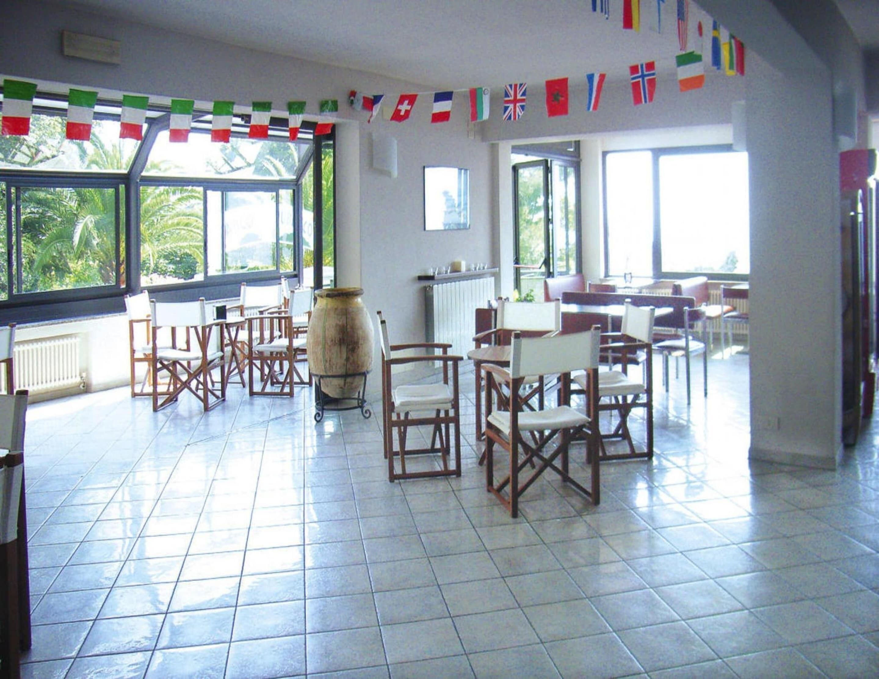 A spacious room with tiled flooring, wooden chairs, and tables. There are large windows allowing natural light, and the room is decorated with international flags.