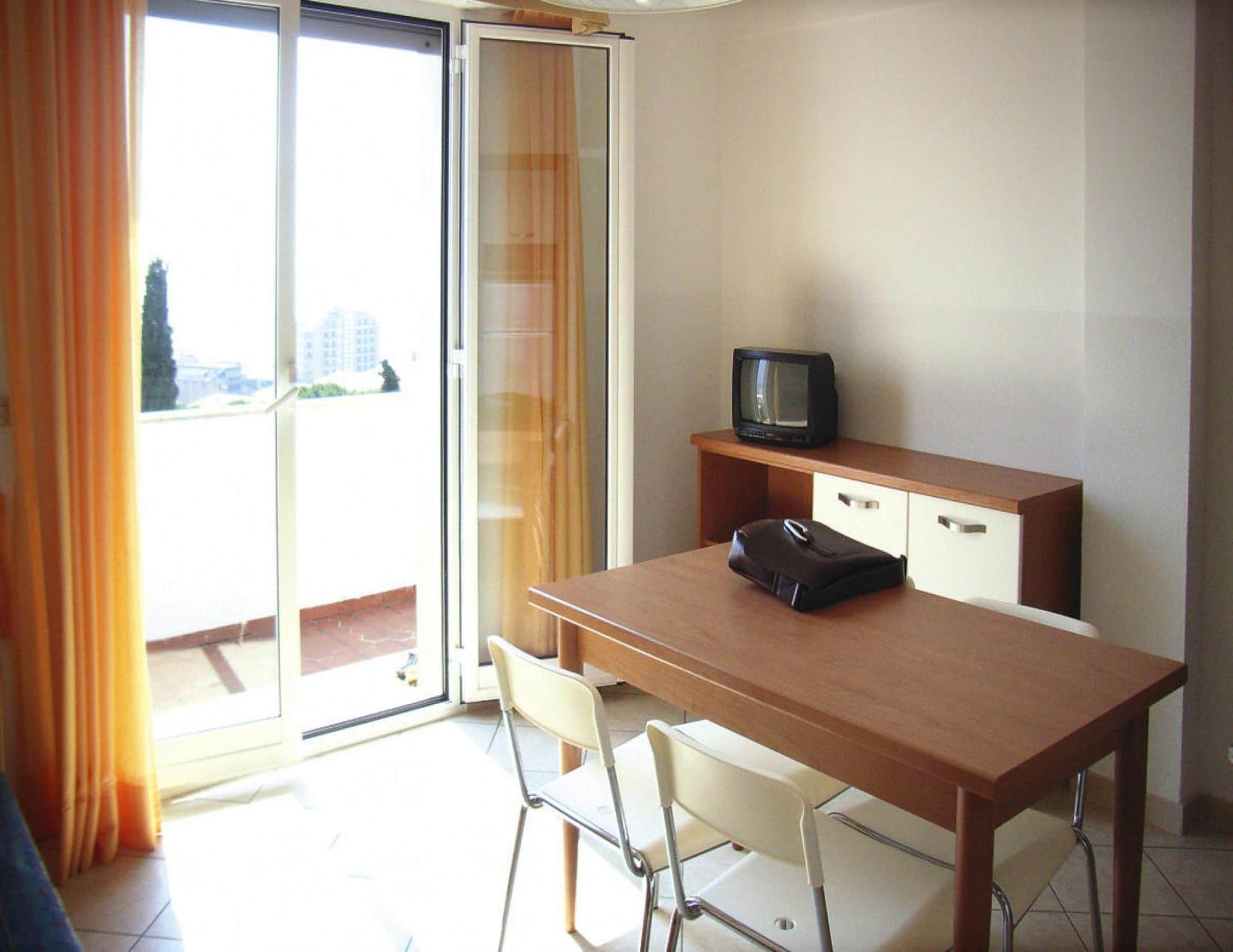 Een kamer met een houten bureau, een televisie, een stoel en een schuifdeur naar een balkon.