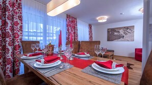 A dining area with a long wooden table set for a meal, red curtains, and a mountain painting on the wall. - Apartment in Neukirchen by Wildkogelbahn (Neukirchen am Grossvenediger)