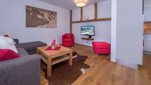 A modern living room with a wooden floor, a red sofa, a grey sofa, a wooden coffee table, and a mounted TV. - Apartment in Neukirchen by Wildkogelbahn (Neukirchen am Grossvenediger)
