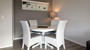 A dining area with a round table and four wicker chairs. There is a white shelf above the table with books and decorative items. A lamp is on the wall to the right. - Ferienwohnung Nahe Nordsee Wattwanderung (Berumbur)