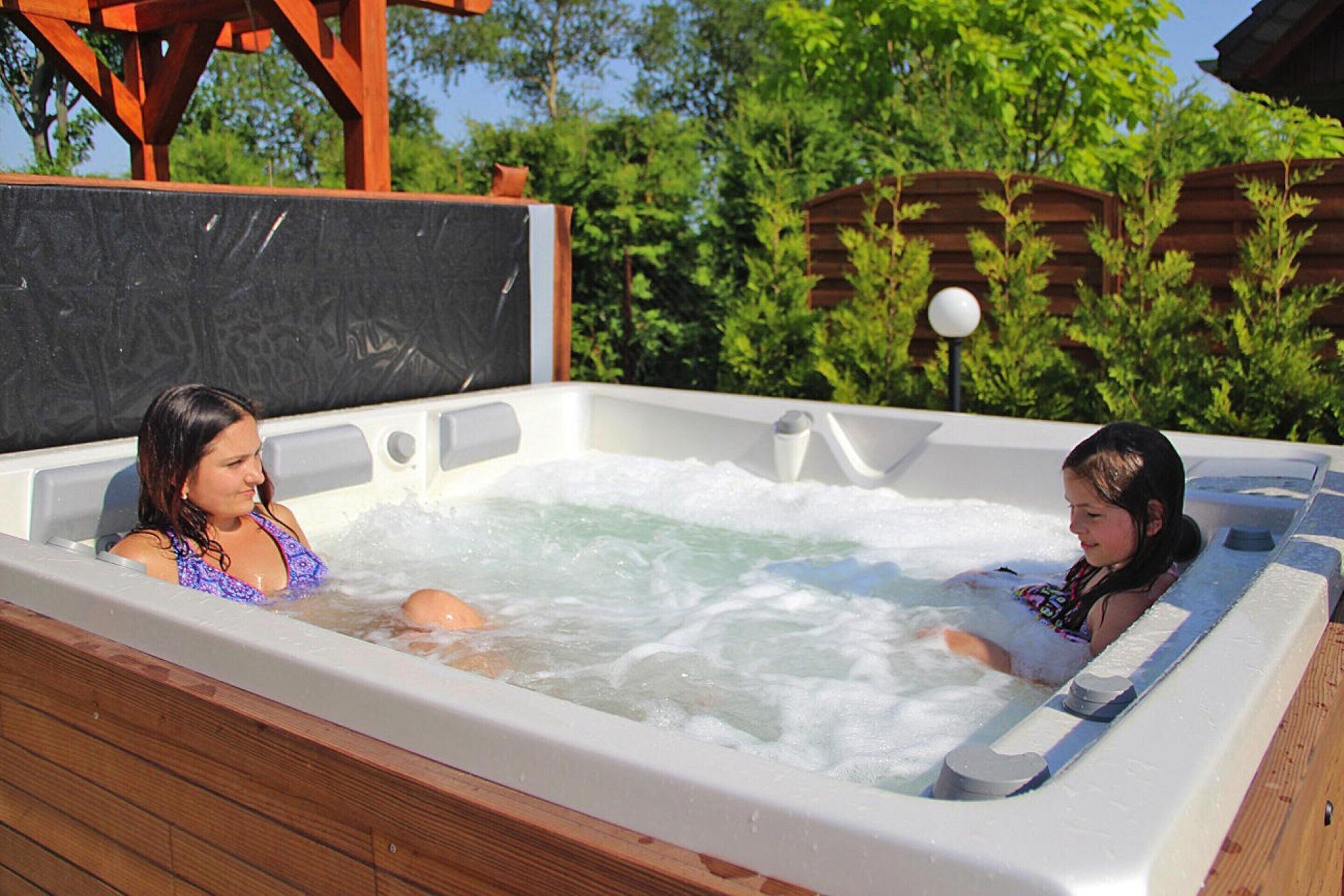 Un bain à remous où se trouvent deux personnes, entouré d’une terrasse en bois et de verdure.