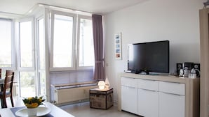 A modern living room with a television, a small table with a vase of flowers, a radiator, and a window with curtains. - Ferienwohnung in Cuxhaven-duhnen (Cuxhaven)